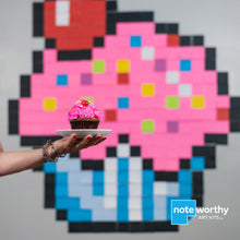 Load image into Gallery viewer, woman holding pink cupcake in front of sticky note pixel art mural