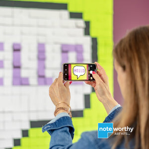 woman taking photo of post it note pixel art on wall with smartphone